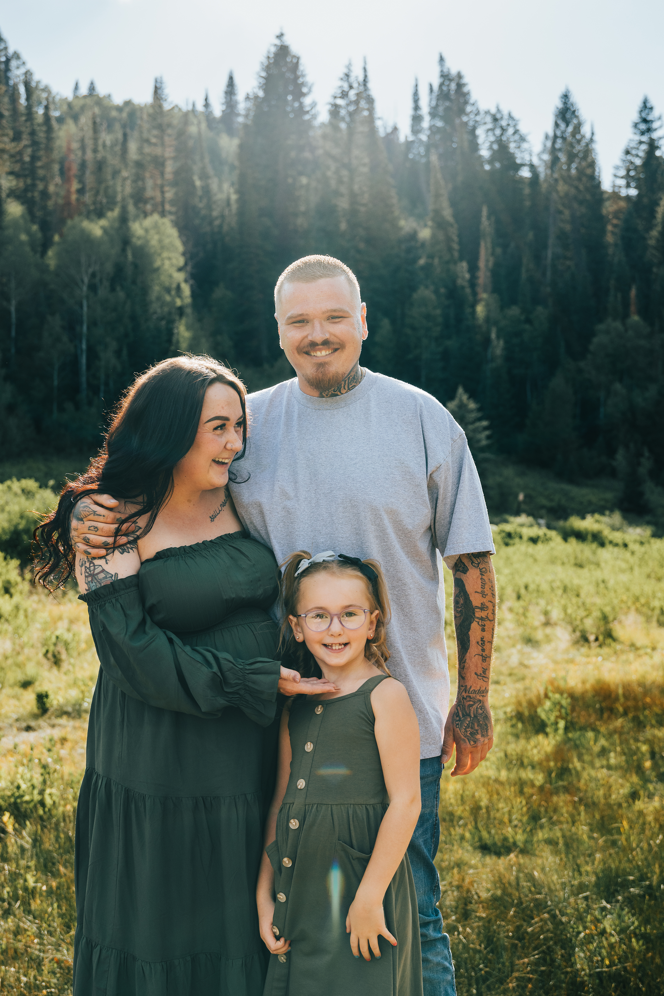 A family of four outdoors in a field with trees in the background. The mother has long dark hair, is wearing a green dress, and is holding a young girl with glasses and a green dress. The father has short blonde hair, tattoos on his arms and neck, an