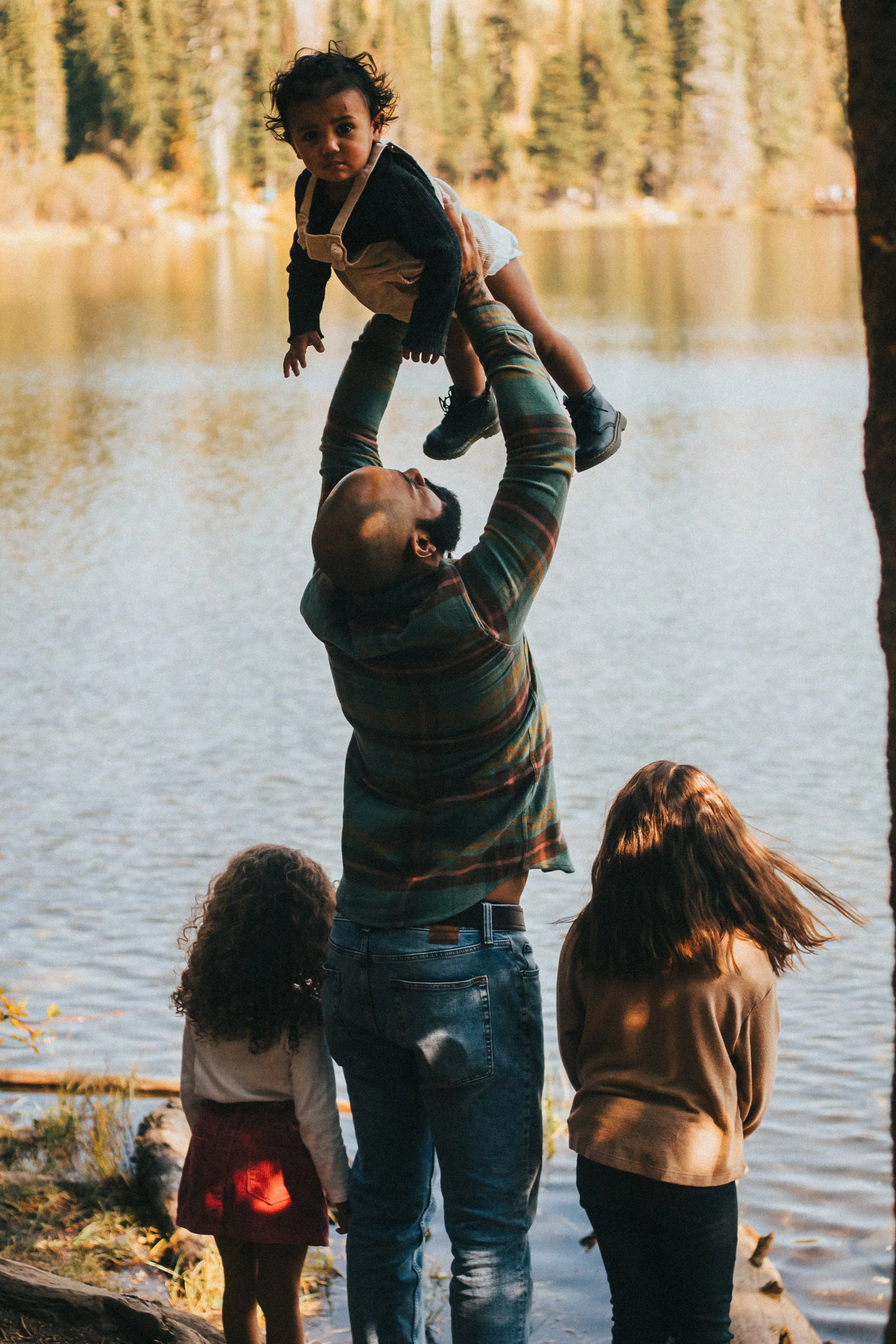 A man lifting a young child in the air near a body of water with trees in the background. Three women with curly hair are facing the water, with their backs to the camera.