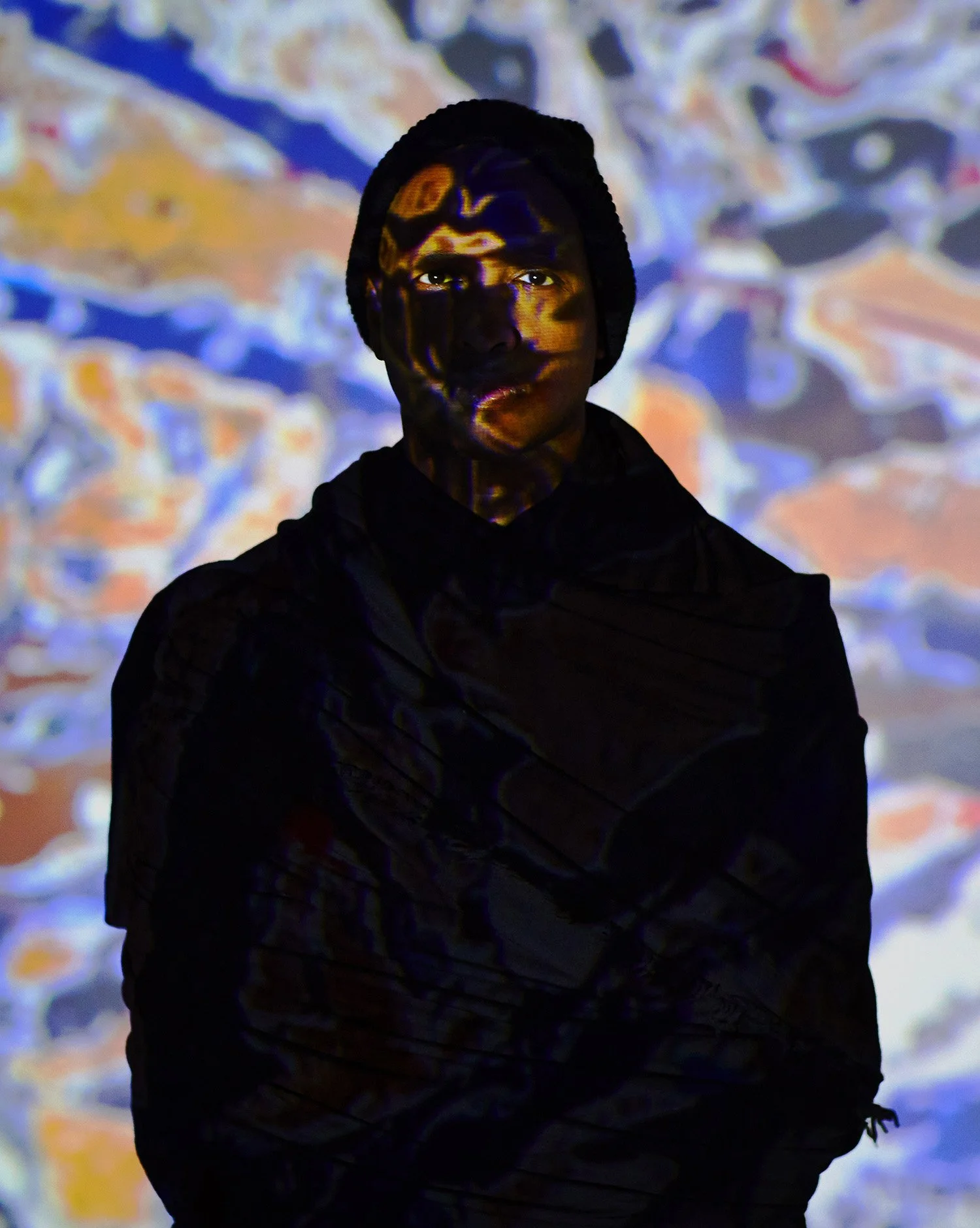 A person with dark skin wearing a black beanie and black jacket, standing in front of a colorful abstract background with projected patterns and faces.