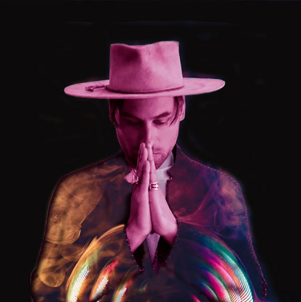 A person wearing a wide-brimmed beige hat, with eyes closed and hands pressed together in a prayer-like pose, against a dark background with colorful light effects.