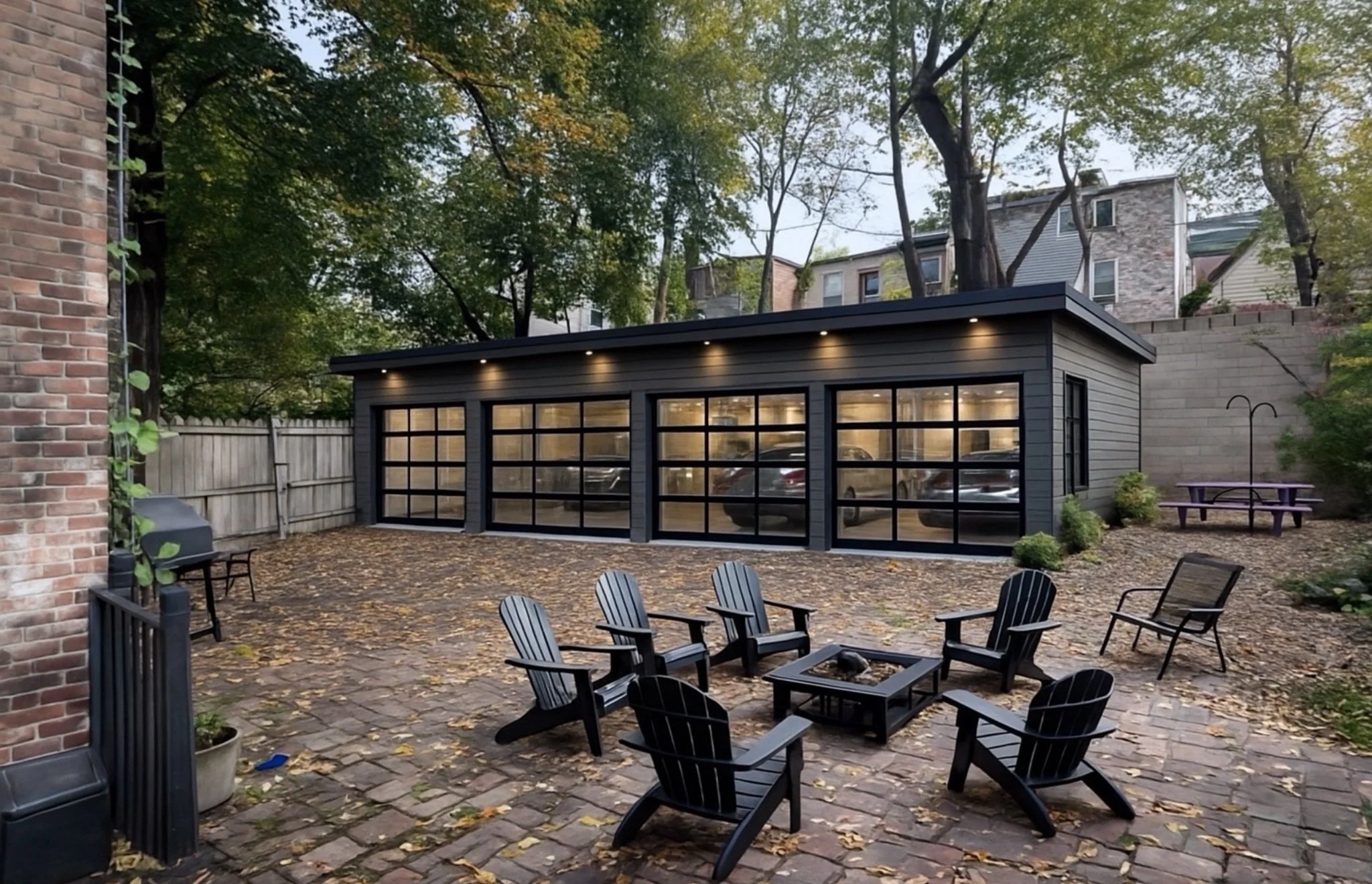 Backyard patio with black Adirondack chairs, brick paved ground, small potted plants, and a modern outside garage with black framing and glass doors. Trees with leaves and a wooden fence are in the background, and some autumn leaves are scattered on the ground.