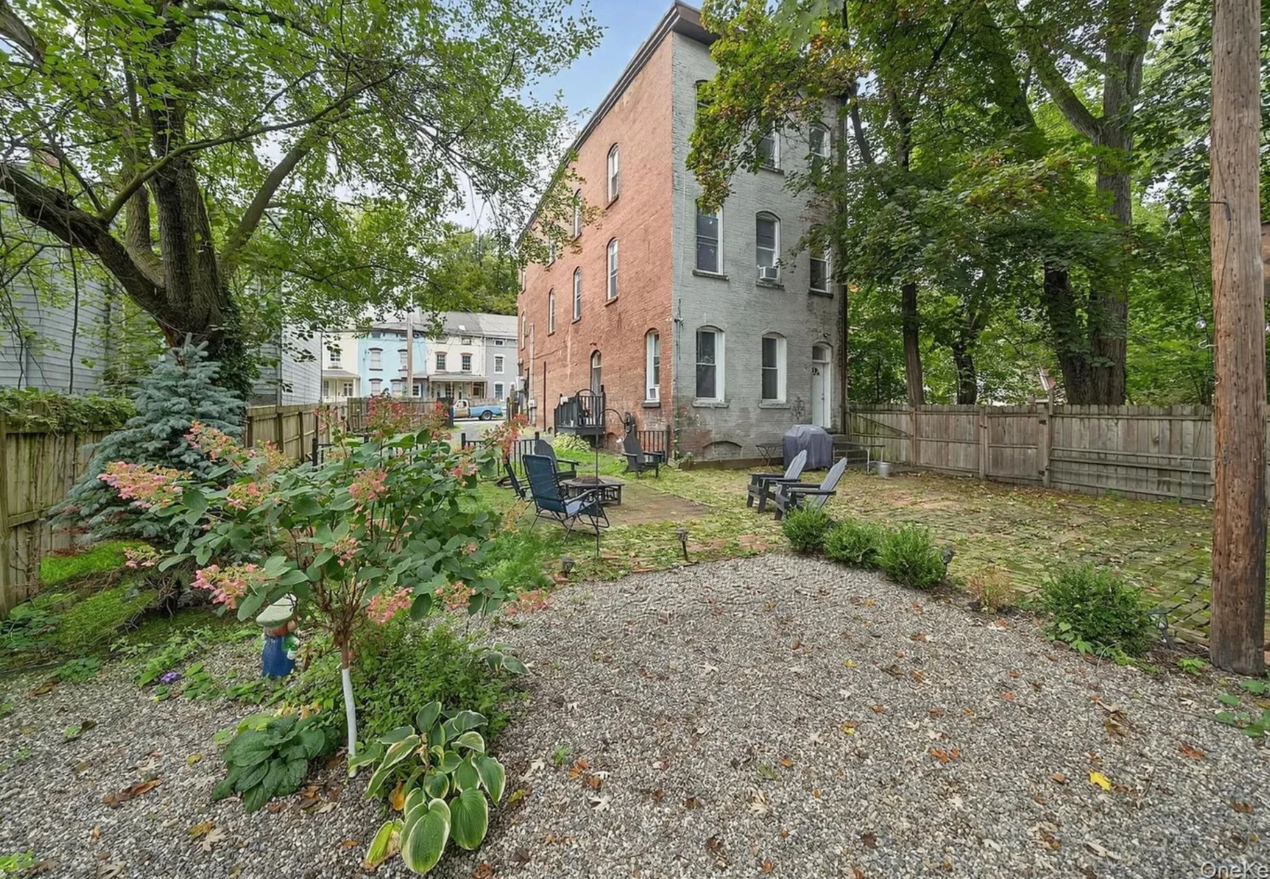 Backyard with chairs, planted bushes, trees, a brick house, and a wooden fence, in a suburban neighborhood.