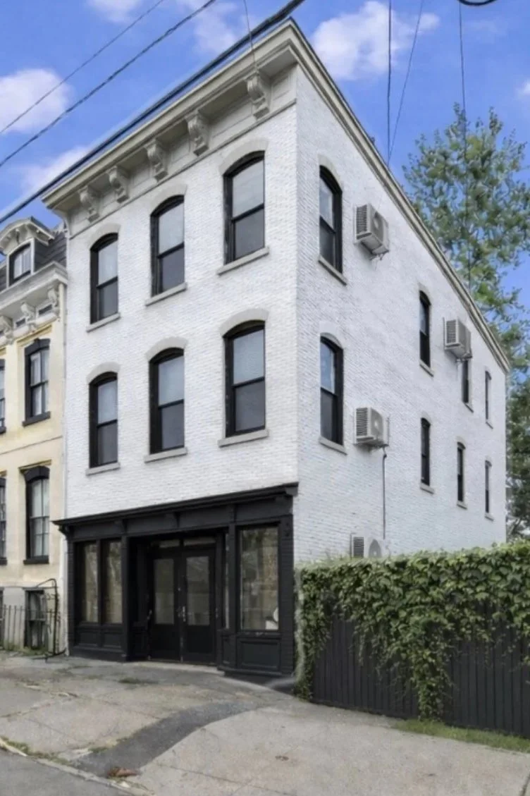 A four-story white brick building with black window trim and air conditioning units attached outside. The building has a black storefront on the ground level and is located on a street with a sidewalk and some greenery.