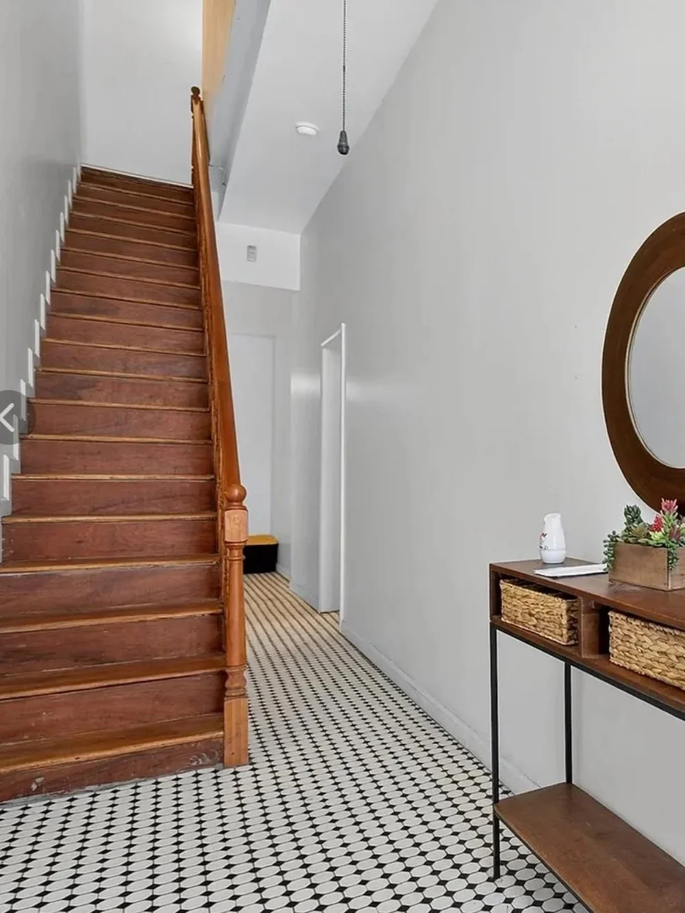 Wood staircase with a wooden handrail in a home, adjacent to a tiled black and white floor, with white walls and a small console table decorated with a plant and other items.