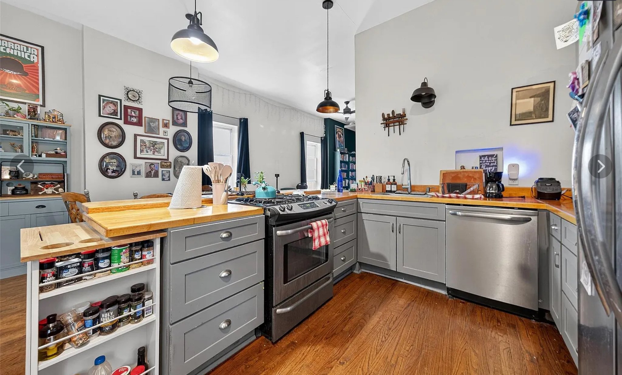 A kitchen with gray cabinets, a wooden countertop, and a white wall decorated with framed photographs and artwork. There is an island with a spice rack, a gas stove, and a sink. Various small appliances and kitchen items are on the countertop, with dark curtains on the windows and pendant lights hanging from the ceiling.