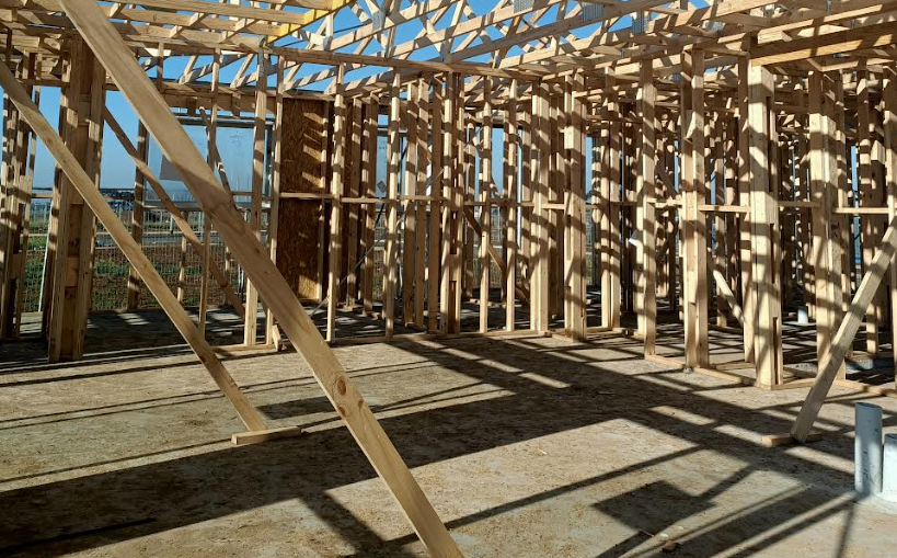 Construction site of a wooden building frame with sunlight casting shadows on the ground.