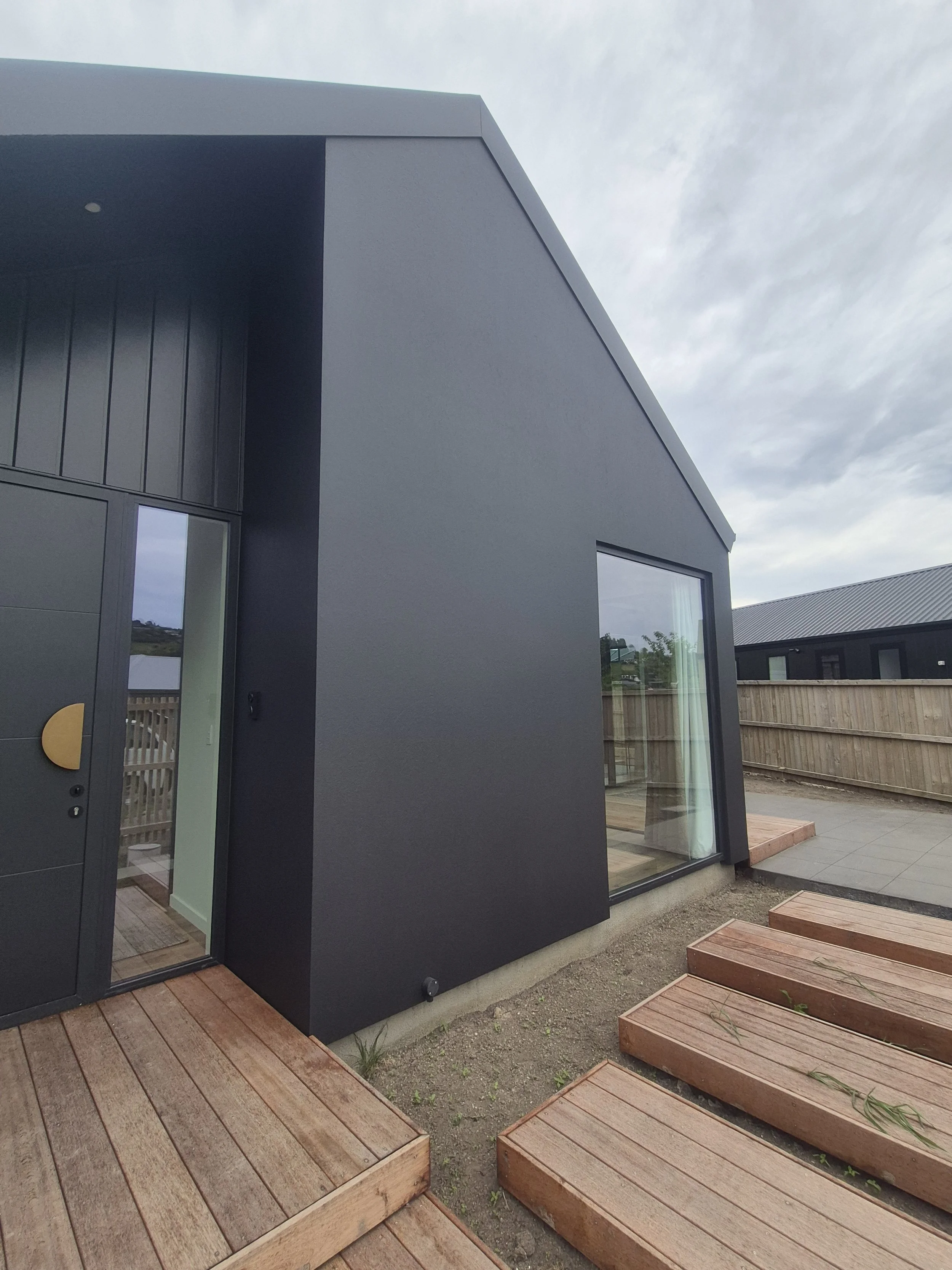 Modern gray house with large glass windows and wooden deck outside, surrounded by a wooden fence and a cloudy sky.