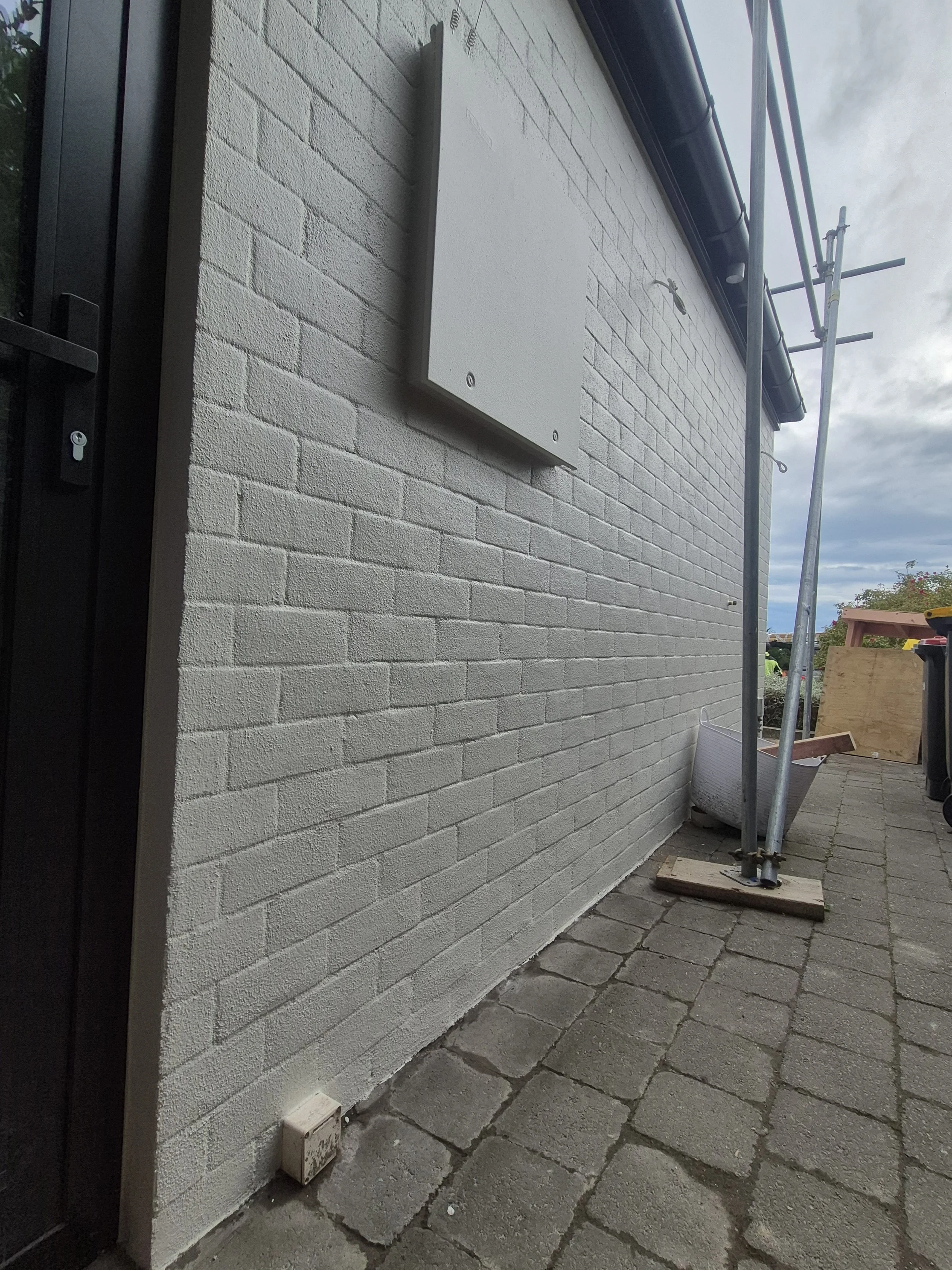 A brick exterior wall of a building with a gray utility box mounted on it, a black door with a lock, and scaffolding set up outside. The ground is paved with stone tiles, and there is a wooden board propped against the scaffolding.
