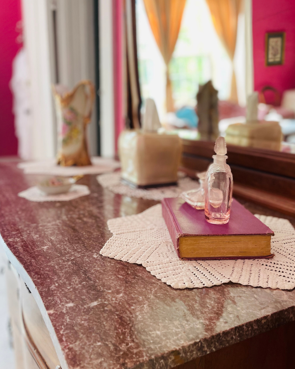 A pink marbled top antique dresser with book, small perfume bottle , tissues and vase set on top. Dresser mirror shows yellow curtains at the front window.