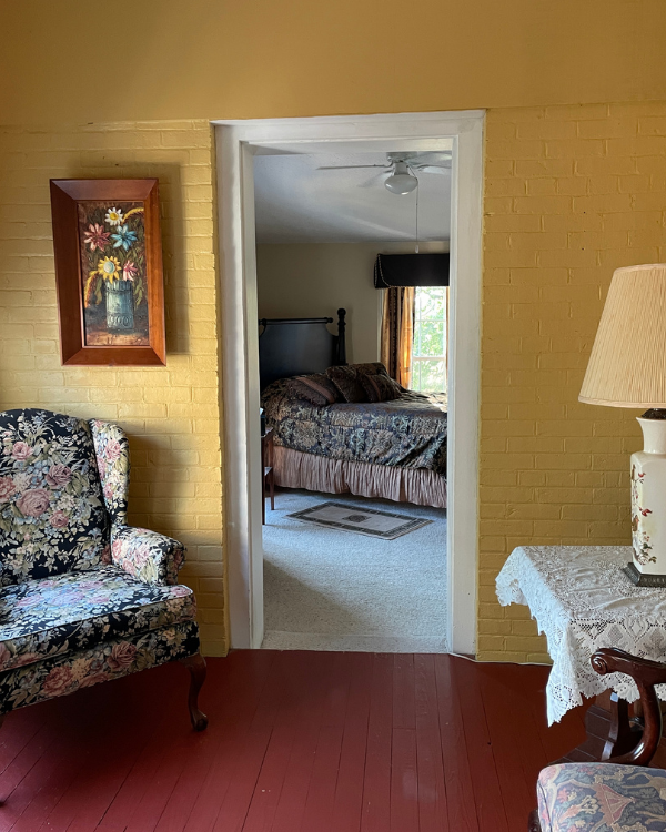 A sunporch with yellow walls and red floor. Floral print winged back chair with framed floral picture above and side table with lamp across. Queen size with bed with black floral print bedspread and pillows in background.