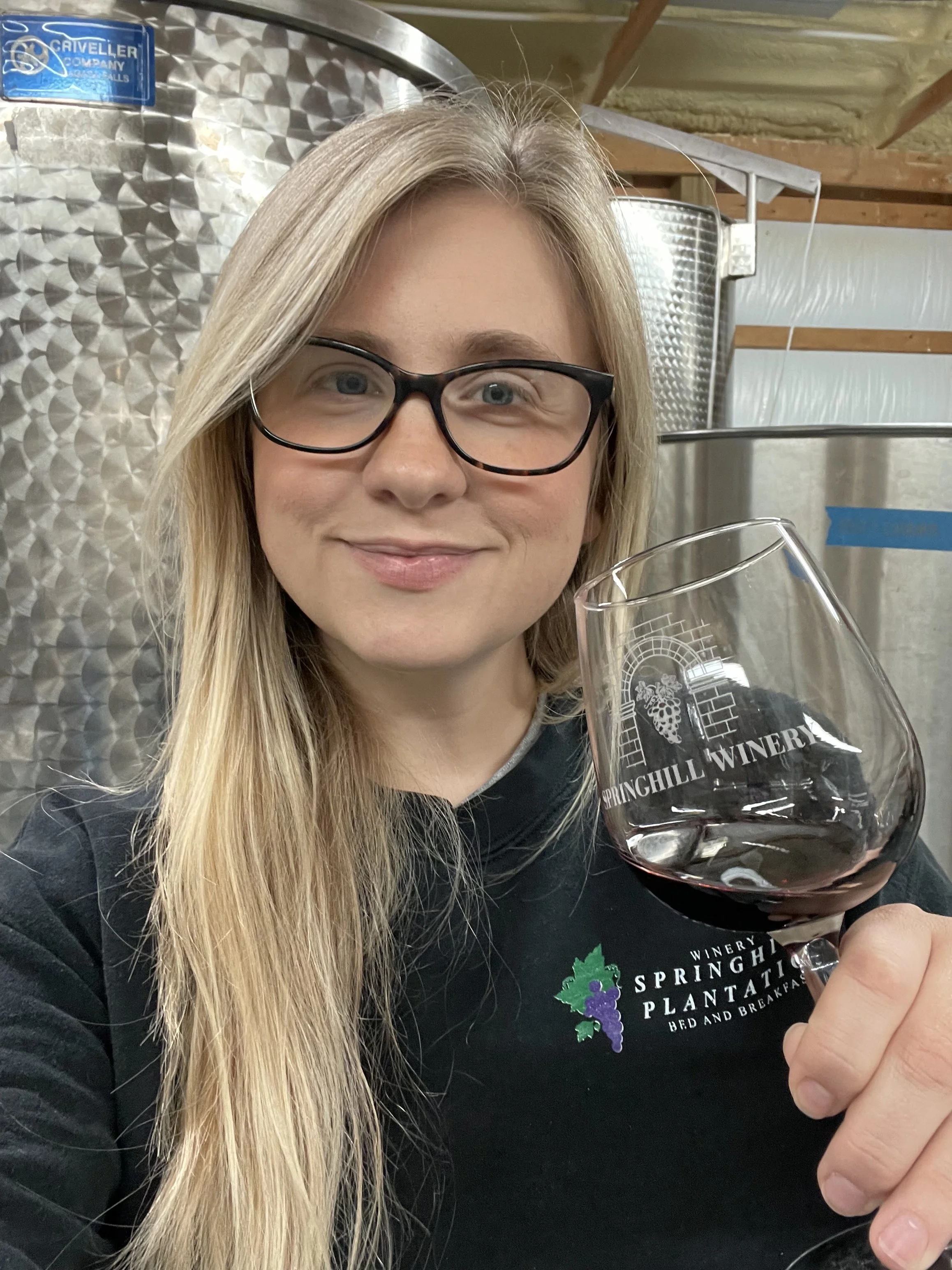 Mallory holding a Springhill Winery glass of red wine in the production room with stainless steel tanks as part of the Springhill winemaking experience.