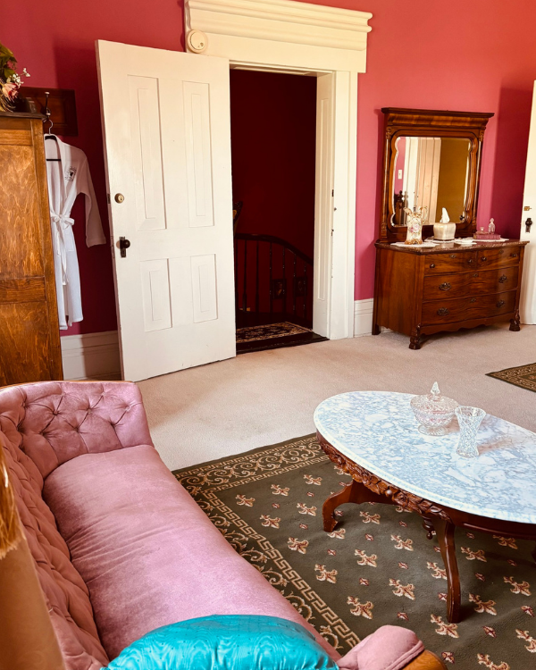 Vintage Rose Suite sitting area featuring a pink velvet sofa, marble‑top table, rose walls, antique dresser, and a robe near the doorway.