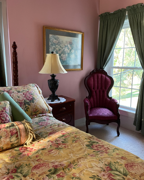 Cozy vintage bedroom with pink walls, floral bedding, red upholstered chair, and window with green curtains.
