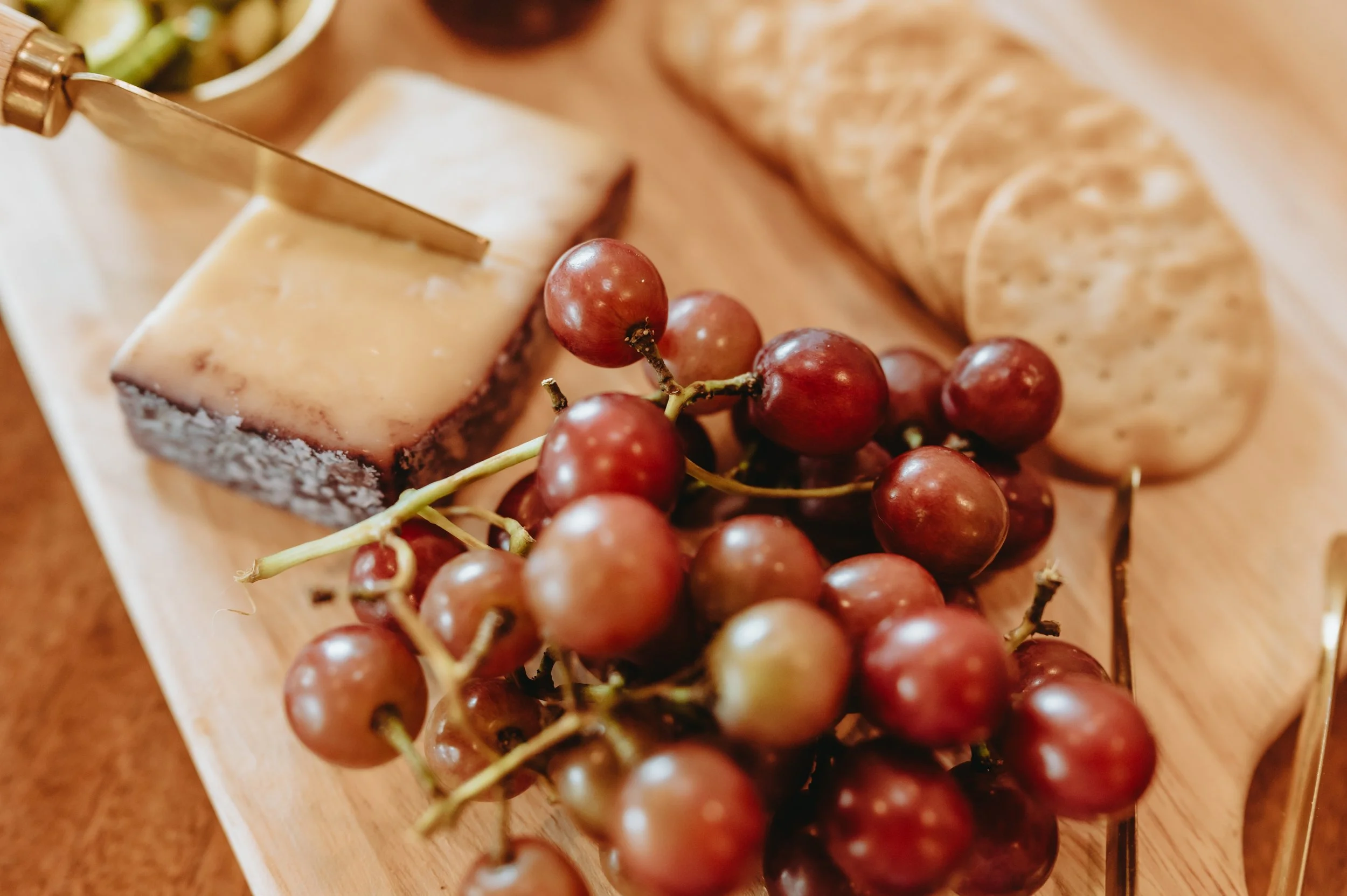 charcuterie board with a knife in a block of cheese, crackers and cluster of grapes.