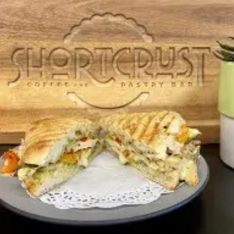 A cutting board with Shortcrust logo and panini sandwich on a doily covered plate