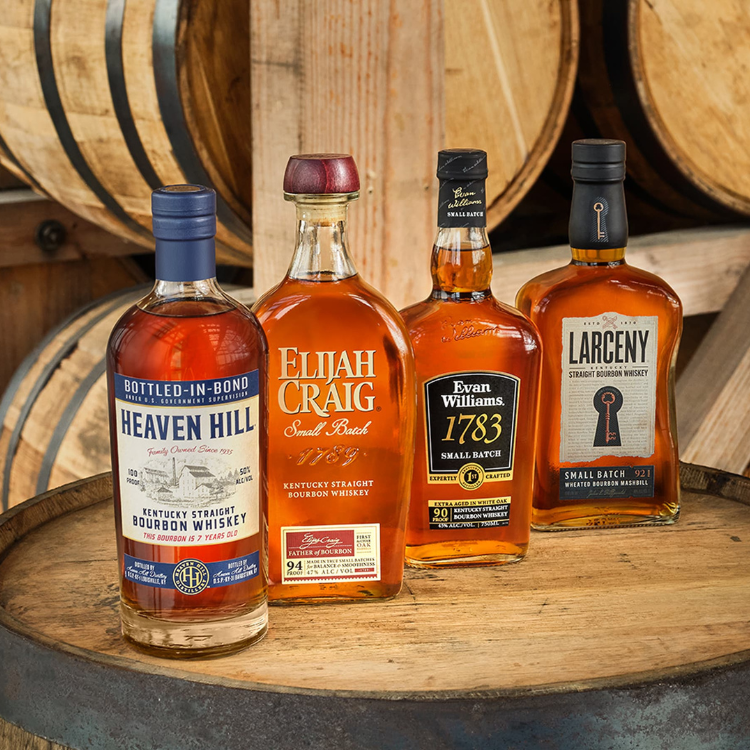 Four bottles of bourbon sit on bourbon barrel with barrels stacked behind.