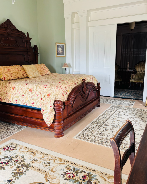 Merlot Suite bedroom with an ornate carved king bed, floral bedding, green walls, antique furnishings, and an adjoining sitting room.
