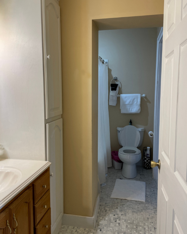 Yellow walled bathroom with white cabinetry. White marbled top oak wood vanity, tiled floor. Toilet, shower curtain and towel on rack in background.