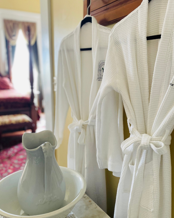 White robes in the Cabernet Suite hanging beside a vintage ceramic pitcher and basin, with the carved bed and red bedding visible behind