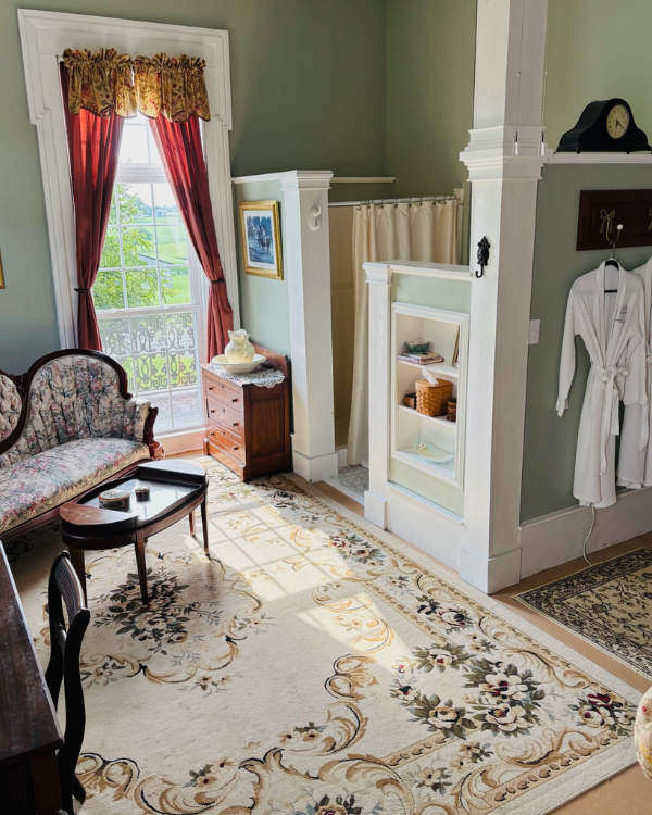 Sitting area in the Merlot Suite with floral sofa, red and gold curtains, antique desk, and a partial bath area with robe and shower.