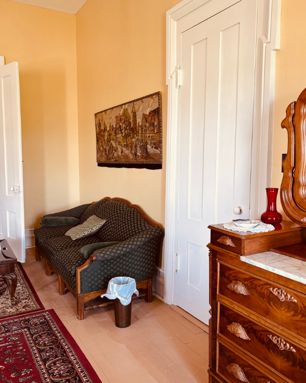 A vintage dresser with a red vase and a tapestry hanging above a patterned sofa in a room with light yellow walls and a red rug.