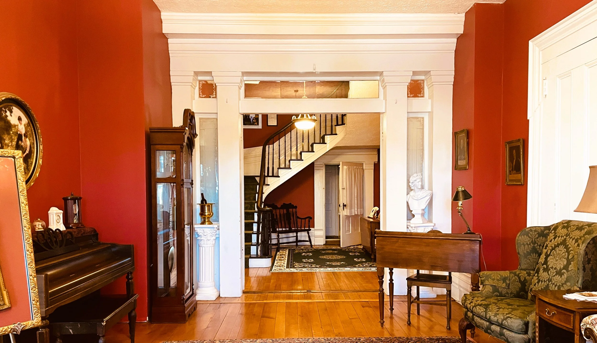 A grand entryway with red walls, a grandfather clock, an antique piano, and a curving staircase visible through an archway.