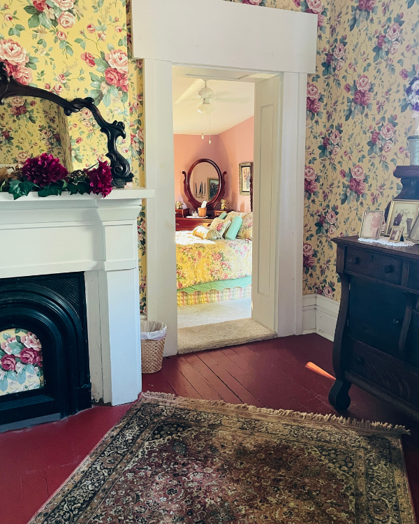A doorway opens to a bedroom with a bed, while the foreground shows a fireplace and a buffet, all decorated with floral wallpaper.
