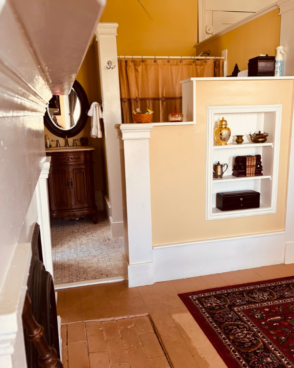 Cabernet Suite featuring a built‑in shelf with décor, warm beige walls, red rug, and a partially open bathroom with vanity and shower.