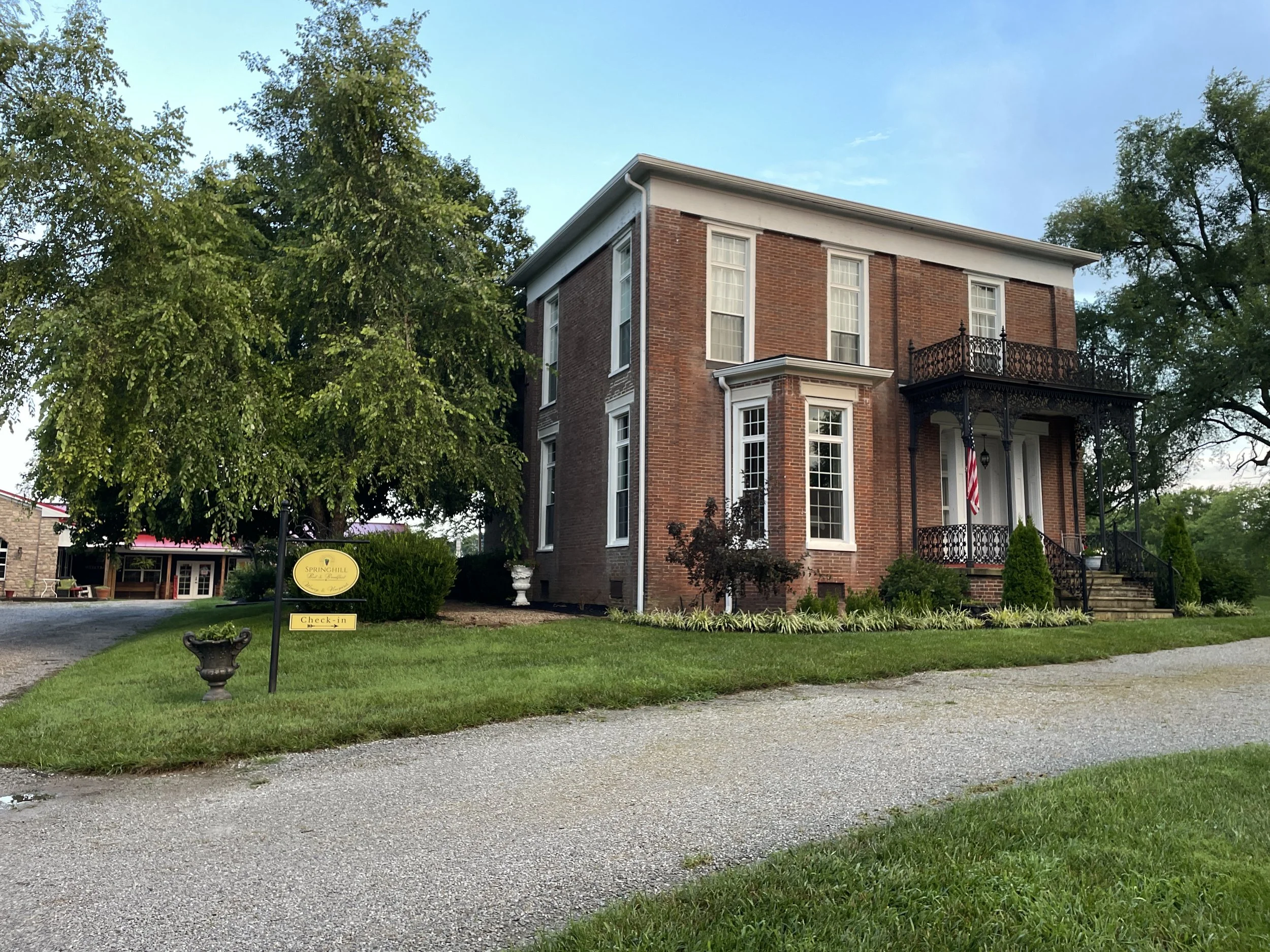 A large two-story brick house with ornate rod iron porch surrounded by trees and landscaping.