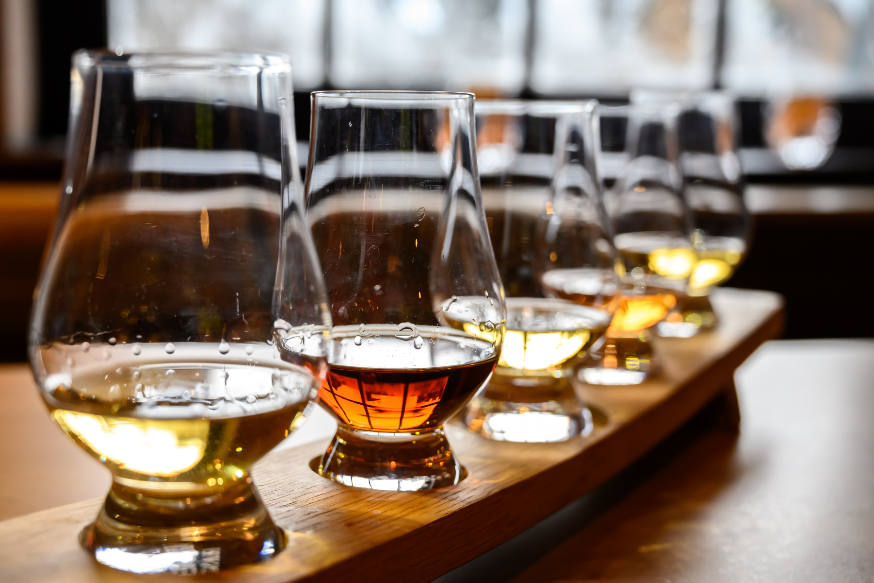 A wooden tasting flight board with seven glasses of different types of wine, from white to red, lined up in a row near a window.
