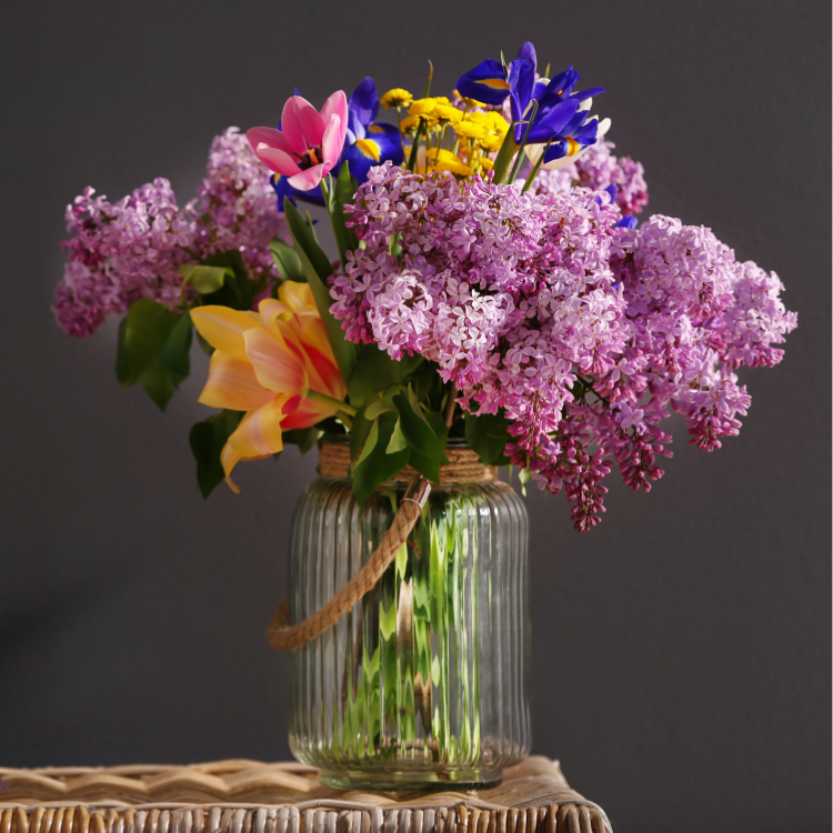 Colorful spring flower bouquet in a ribbed glass vase with lilacs, daisies, tulips, and irises.