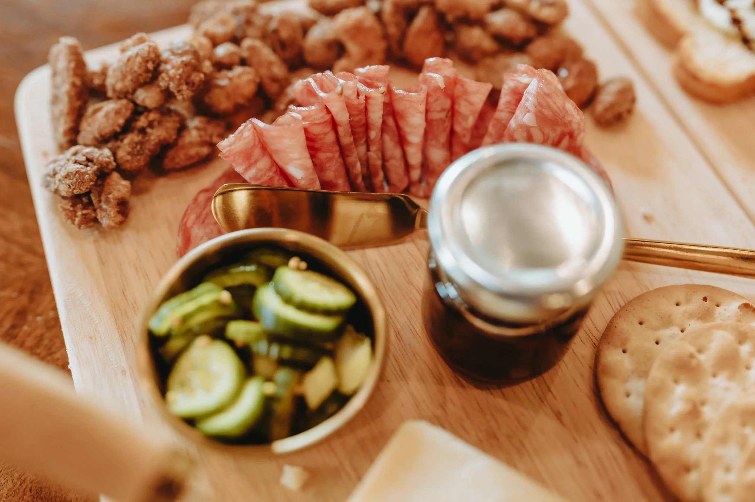 Charcuterie board with a cup of pickles, small jar of jam, crackers, meat and nuts.