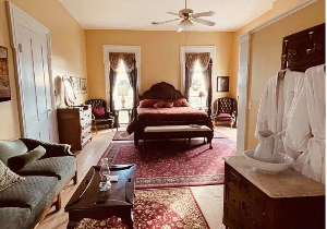 A large spacious room with a four-poster bed, a sofa, a coffee table with a lamp, and several armchairs, with white robes hanging on a rack near a vanity with a pitcher and basin