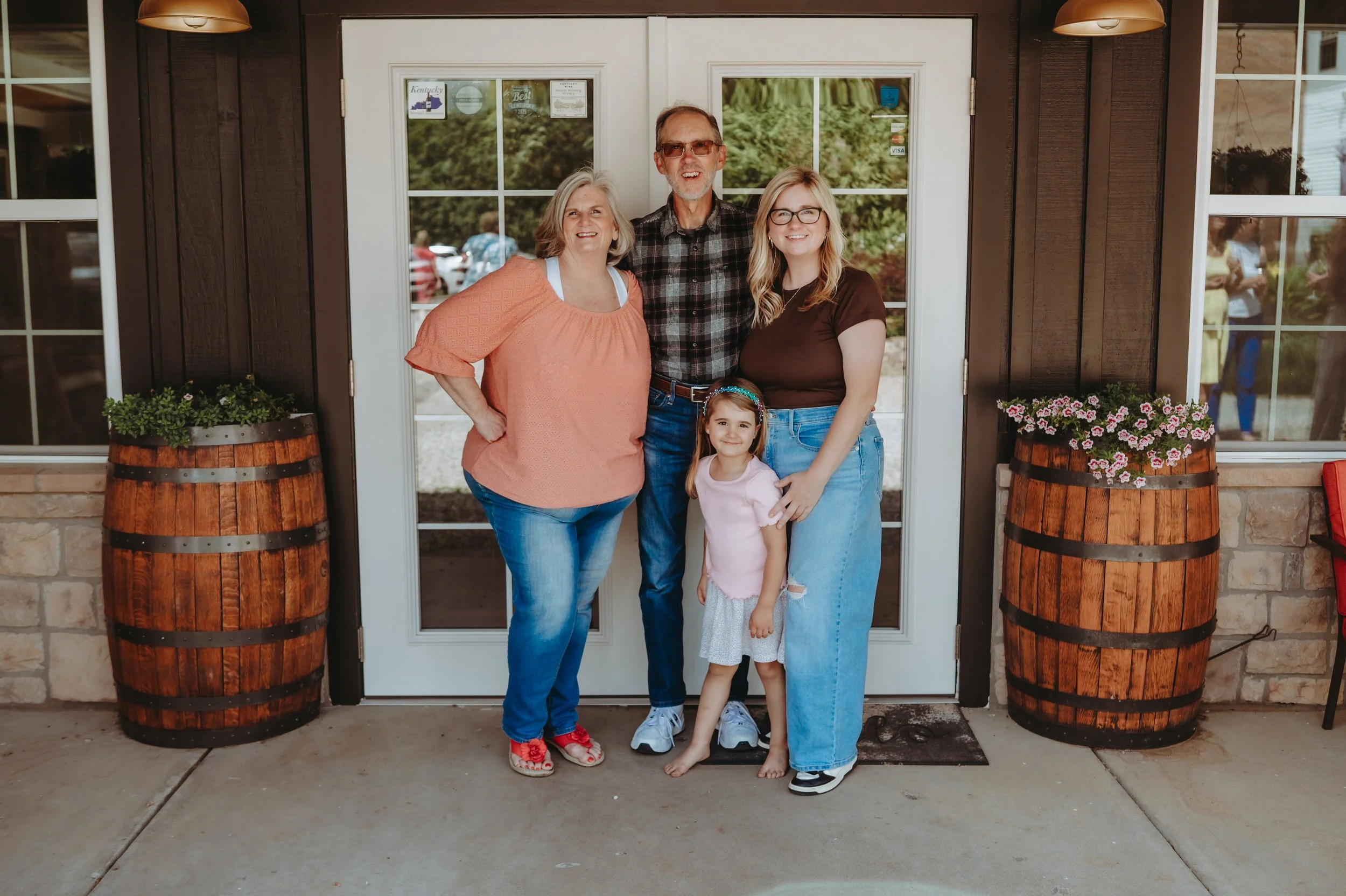 Family standing in front of the Springhill winery doors with flower‑filled bourbon barrels on each side.