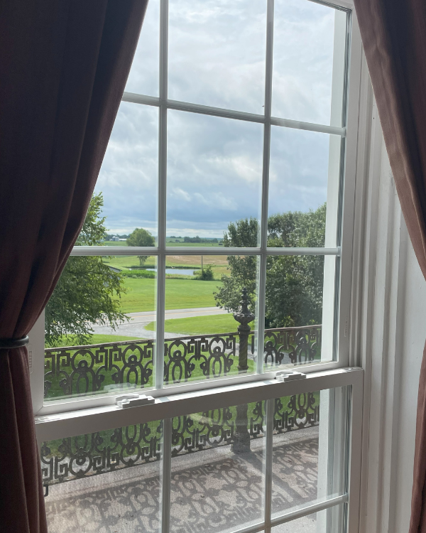 View of a green landscape with trees and a body of water through a window with white mullions and dark curtains.