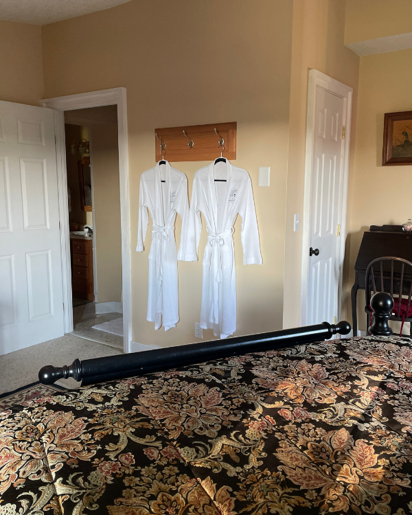 White robes hang on a cream-colored wall. Doorway leads into bathroom to the left. White door on the right. A chair sits in front of a antique desk with framed picture above. Black floral print comforter in foreground.