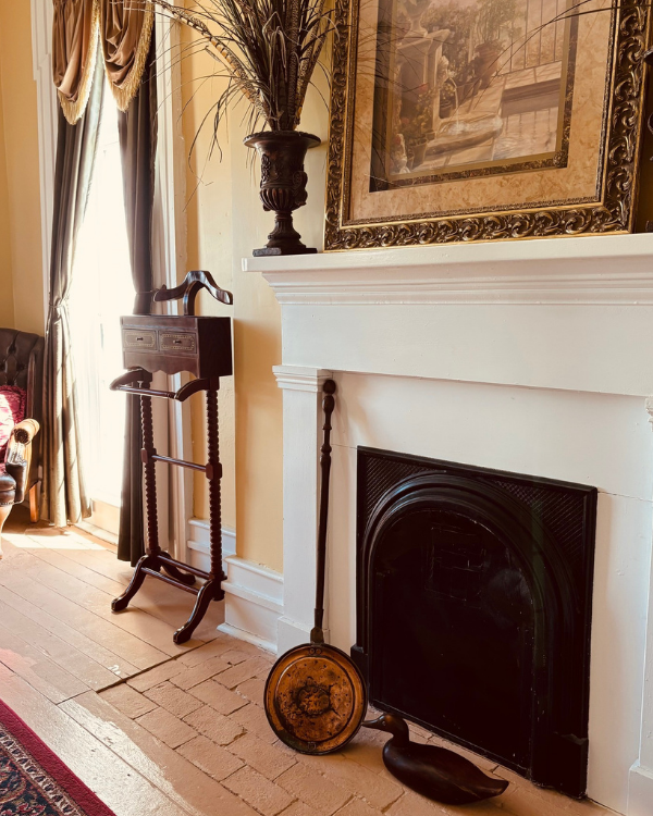 A vintage wooden valet stand with drawers and a coat rack is placed next to a fireplace with a decorative fire screen and a wooden duck decoy.
