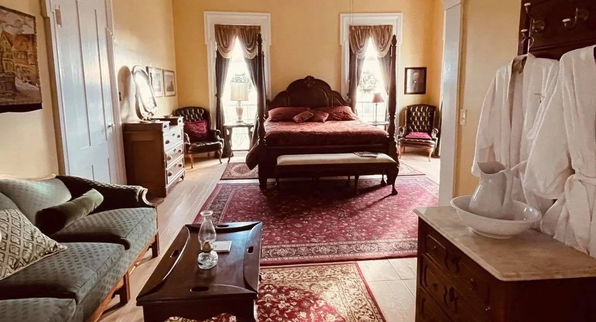 A large spacious room with a four-poster bed, a sofa, a coffee table with a lamp, and several armchairs, with white robes hanging on a rack near a washstand with a pitcher and basin