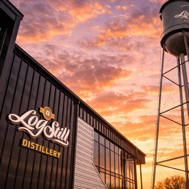 Exterior of Log Still Distillery in Gethsemane, Kentucky, featuring a modern metal‑sided building and branded water tower set against a vibrant sunset sky.