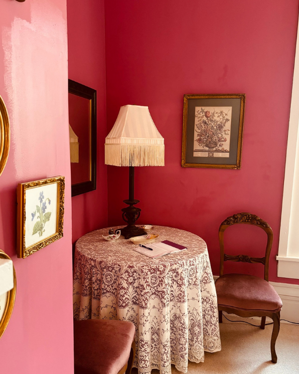 A corner nook of pink walls with a lace tablecloth covering a table. A lamp and book set on the table with a chair two chairs beside the table. A floral picture hangs on the wall.