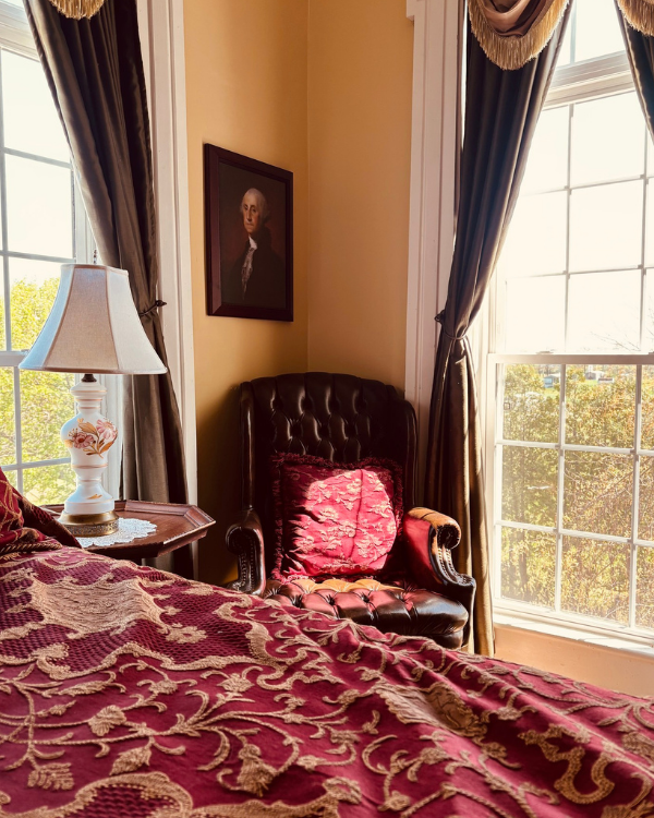 Cozy Cabernet Suite corner featuring a tufted leather chair, elegant drapery, floral lamp, and richly patterned bedding.