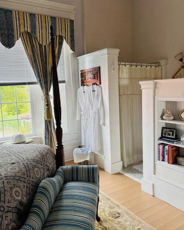 A striped chaise lounge sits at the foot of a four-poster bed, with striped curtains and valance in background. White robes are hanging on the wall. Shower curtain is seen in the bathroom. Corner post with open shelves to the right with books.