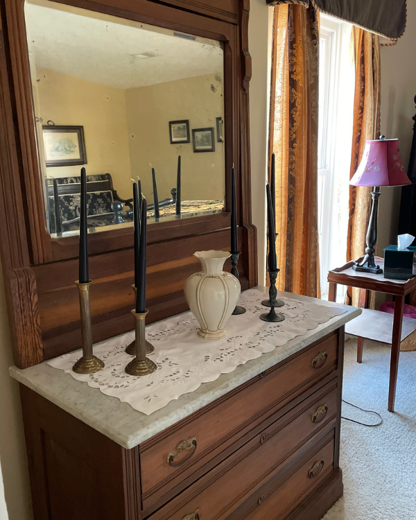 Antique marble top dresser with mirror. A vase and candlesticks sit on top of laced doily. Side table, with lamp and bed. Striped curtain and valance at window.
