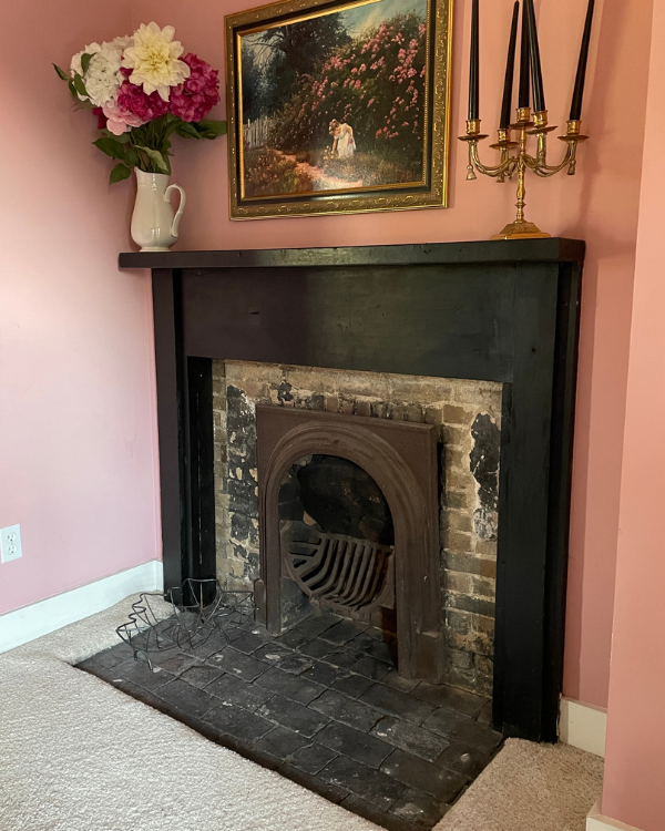 Vintage corner fireplace with dark wooden mantel, floral arrangement, black candles, and framed pastoral painting in a pink room.
