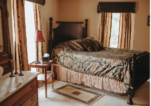 A queen size bed with black floral comforter and pillows set in the corner of bedroom with side table and lamp. Antique dresser with marble top and mirror in foreground to the left with candlesticks set on top.