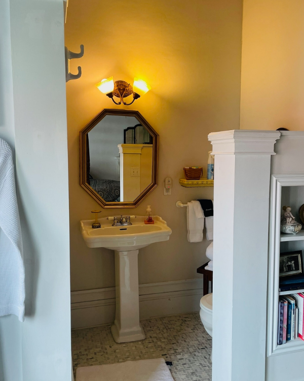A pedestal sink with framed mirror and light above. Corner post and open shelves to the right with books.