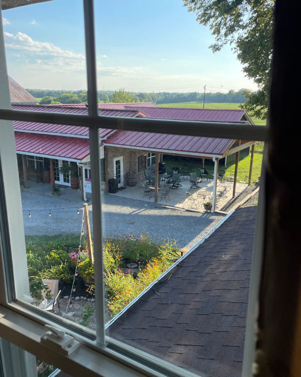 A view out a window of a red roofed building with covered patio seating. A gravel drive lines a flower garden.