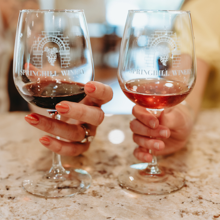 Two hands holding Springhill Winery glasses filled with red and rosé wine on a countertop.