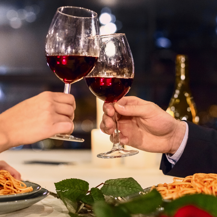 Two hands clinking red wine glasses over a candlelit dinner with pasta and roses.