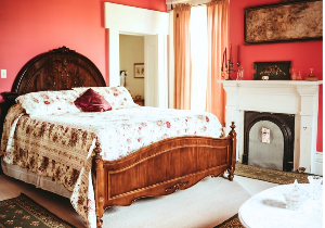 A bedroom features a large wooden bed with a floral comforter and a burgundy accent pillow, set against a pink wall. A white fireplace with a decorative mantle is visible to the right.